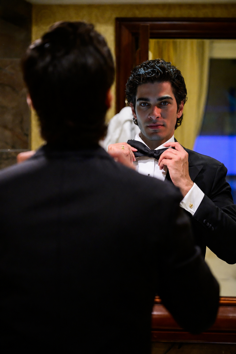 De La Plata adjusting his bow tie in a mirror, wearing a tuxedo